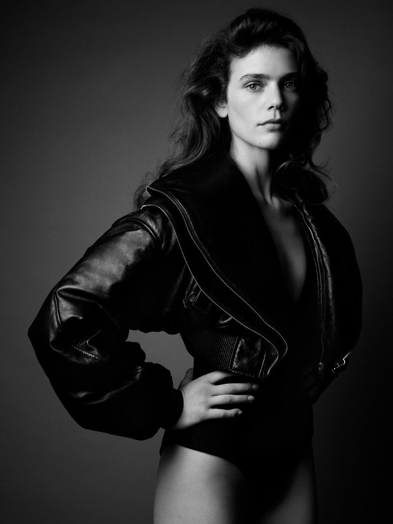 Black and white studio portrait of a woman with wavy hair, wearing a black jacket and bodysuit against a gray background.