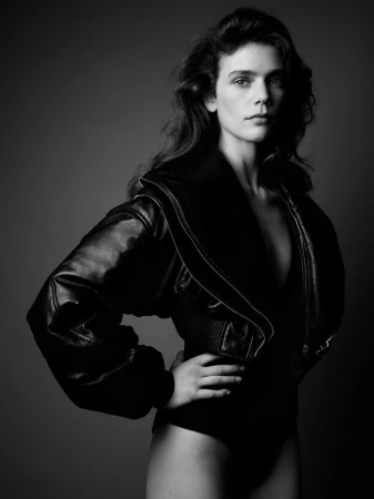 Black and white studio portrait of a woman with wavy hair, wearing a black jacket and bodysuit against a gray background.