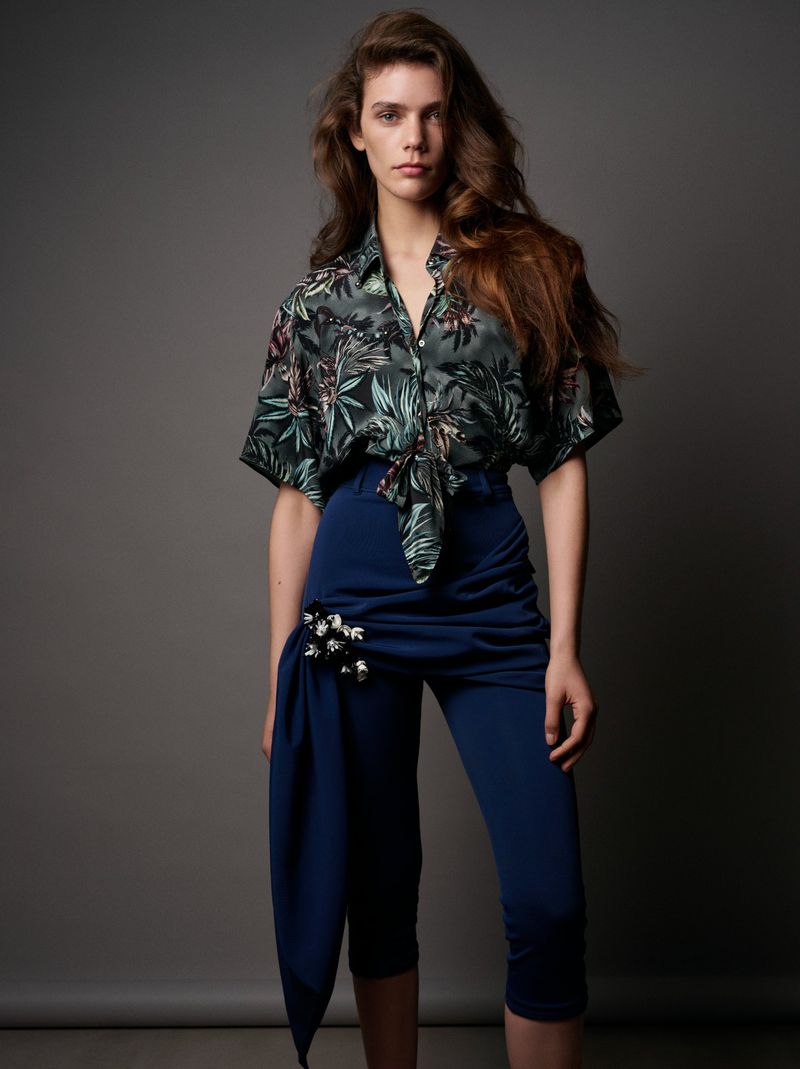 Fashionable woman in floral shirt and capri pants poses in a studio, showcasing contemporary style and apparel in portrait.
