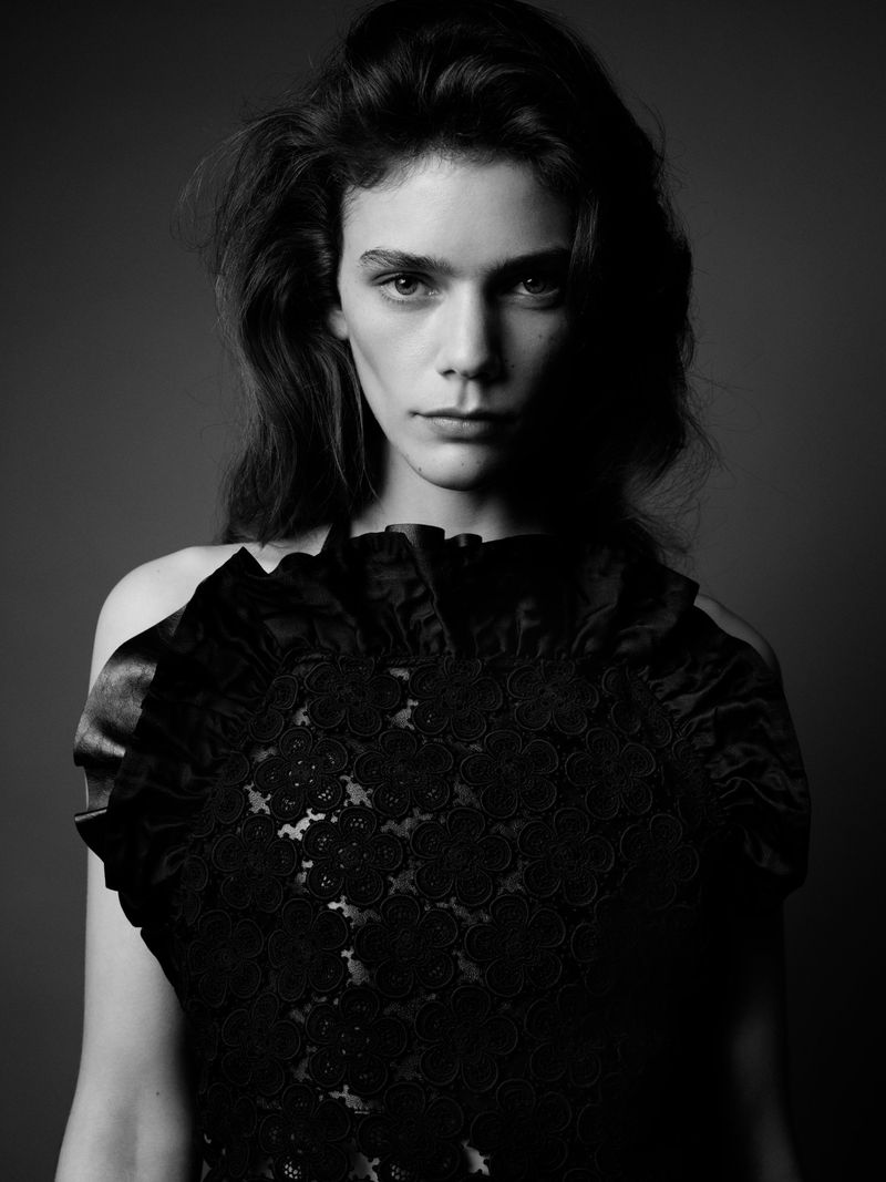 Monochrome portrait of a woman with long wavy hair wearing a ruffled black top against a dark gray background in a photo studio.