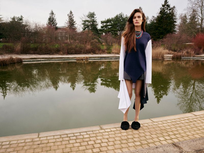 A model poses in an elegant navy and white color-block dress near a pond surrounded by trees in a serene outdoor setting on an overcast day.