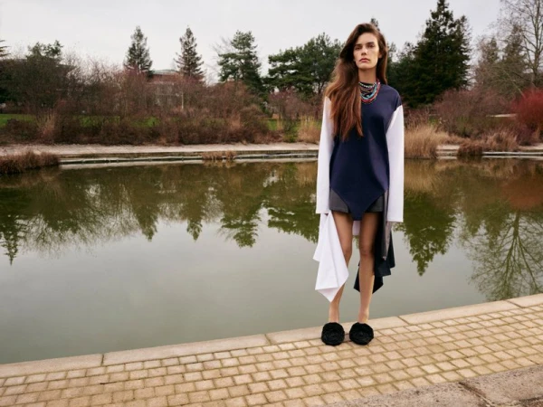 A model poses in an elegant navy and white color-block dress near a pond surrounded by trees in a serene outdoor setting on an overcast day.