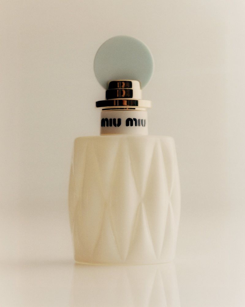 Miu Miu perfume bottle with a white, geometric-patterned body and a light blue round cap against a neutral, minimalist backdrop.