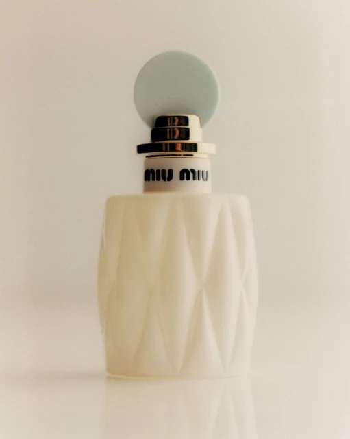 Miu Miu perfume bottle with a white, geometric-patterned body and a light blue round cap against a neutral, minimalist backdrop.