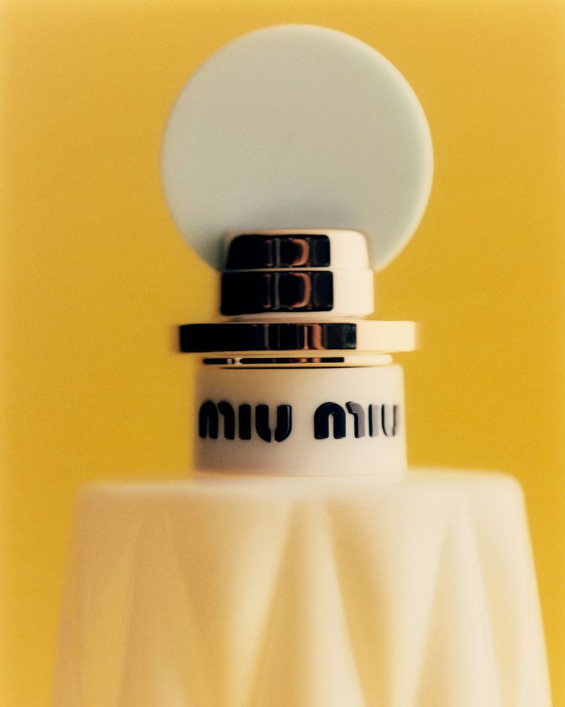 Close-up studio shot of a Miu Miu perfume bottle with light blue cap and textured glass, set against a vibrant yellow backdrop.