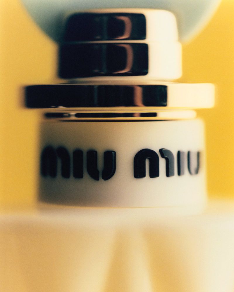 Close-up studio shot of a Miu Miu perfume bottle with a white and black cap, against a soft yellow background creating a minimalist aesthetic.