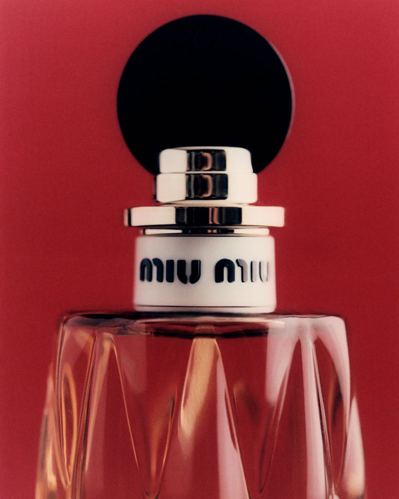 Close-up studio shot of Miu Miu perfume bottle, showcasing its faceted glass design, black topper, and logo against a red backdrop.
