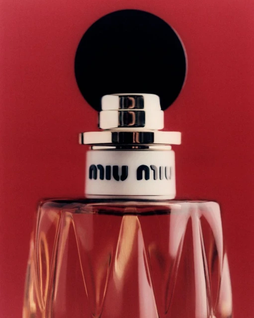 Close-up studio shot of Miu Miu perfume bottle, showcasing its faceted glass design, black topper, and logo against a red backdrop.