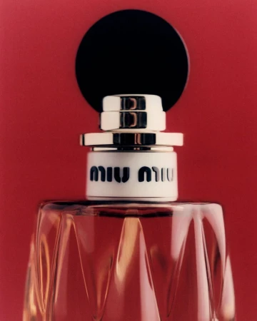 Close-up studio shot of Miu Miu perfume bottle, showcasing its faceted glass design, black topper, and logo against a red backdrop.