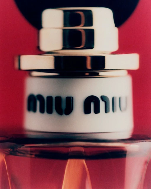 Close-up shot of a Miu Miu perfume bottle with a gold and white cap, set against a red background, showcasing the brand's logo.