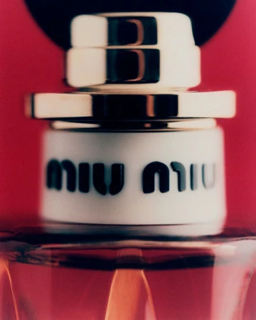 Close-up shot of a Miu Miu perfume bottle with a gold and white cap, set against a red background, showcasing the brand's logo.