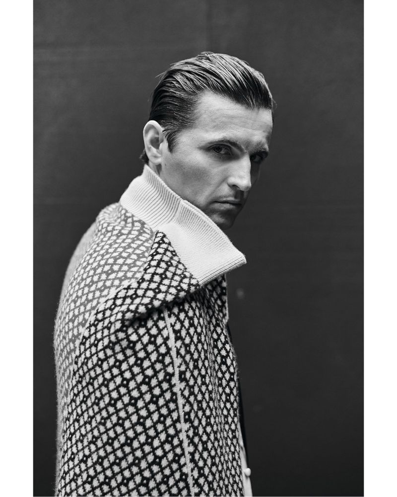 Black and white portrait of a man with slicked-back hair, wearing a patterned coat, looking over his shoulder against a dark background.