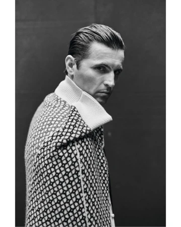 Black and white portrait of a man with slicked-back hair, wearing a patterned coat, looking over his shoulder against a dark background.