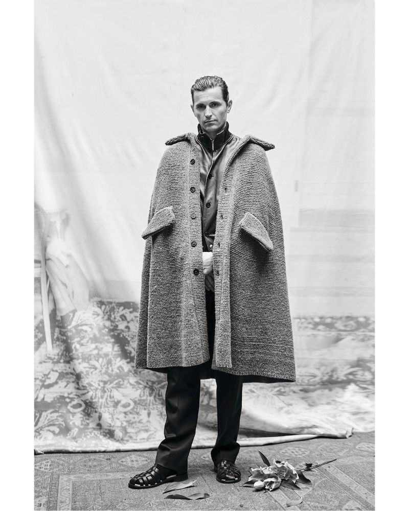 Monochrome portrait of a man in a cape-like coat, dark trousers, and sandals, standing on a patterned rug, against a fabric backdrop.