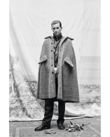 Monochrome portrait of a man in a cape-like coat, dark trousers, and sandals, standing on a patterned rug, against a fabric backdrop.