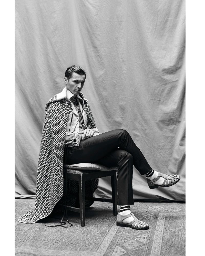 Black-and-white portrait of a man in dark trousers, collared shirt, patterned cape, and sandals sitting on a chair on a patterned rug.