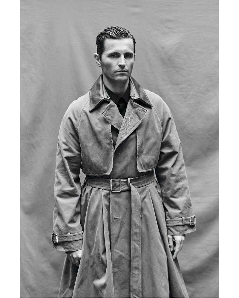 Monochrome portrait of a man in a trench coat. He is centered against a neutral backdrop, and the shot is medium framing.