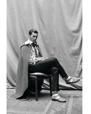 Man seated on a chair wearing a patterned cape and sandals against a draped backdrop in a monochrome portrait.