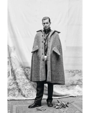 Monochrome portrait of a man wearing a textured wool cape and dark trousers standing before a patterned backdrop.
