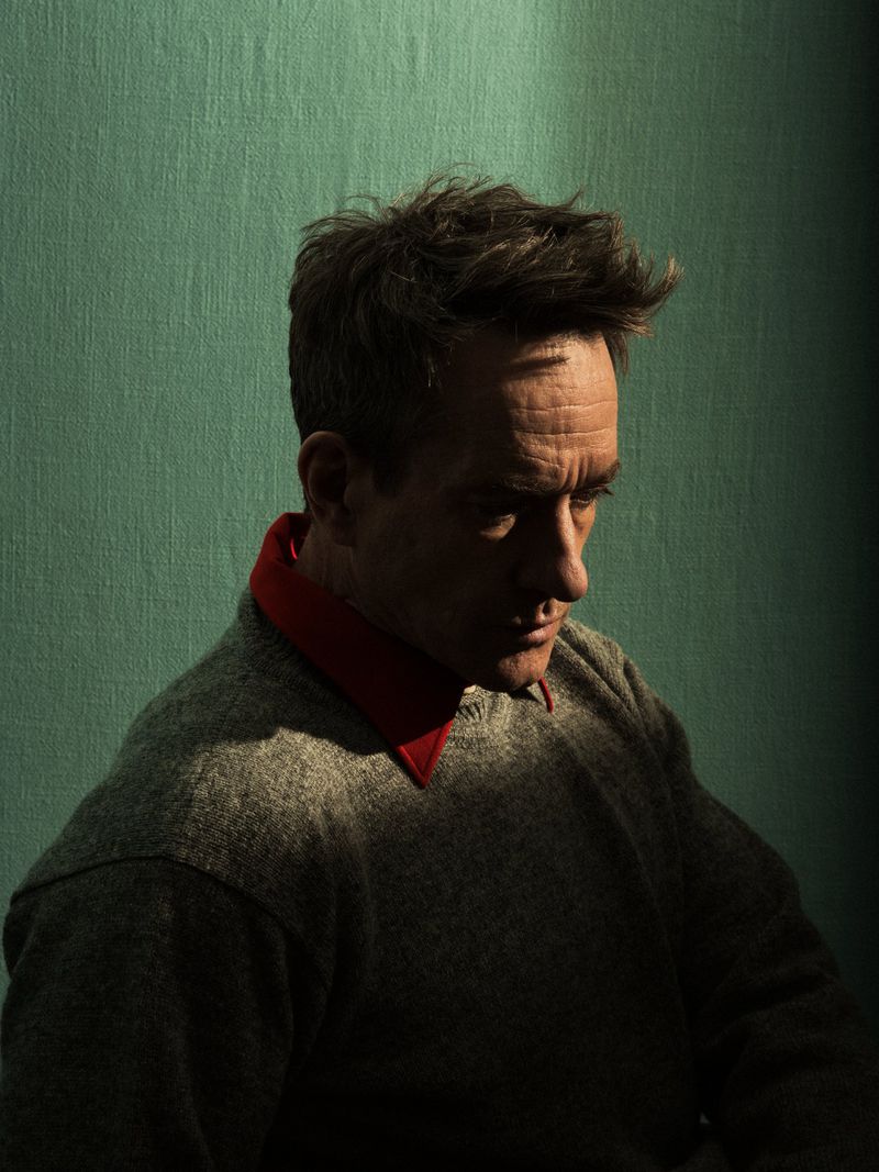Portrait of a light-skinned adult male with brown hair wearing a gray sweater with a red collar, against a green textured wall.