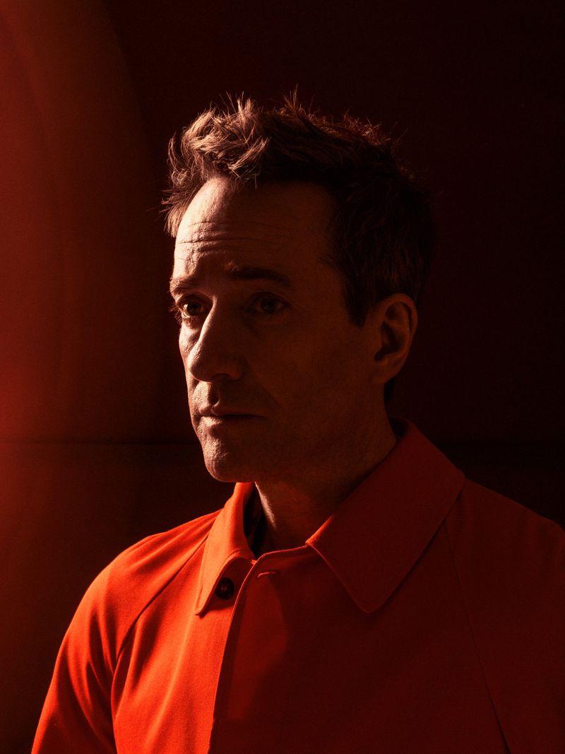 Portrait of an adult male with short hair, wearing an orange collared shirt, against a dark background with warm lighting highlighting his face.