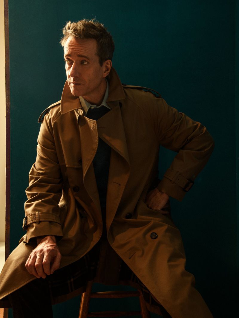 Adult man in tan trench coat seated on wooden stool with a neutral expression against teal backdrop in studio lighting.