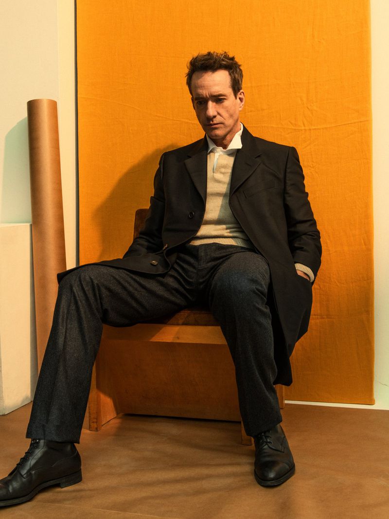 Portrait of an adult male sitting on a chair in a photo studio. He is wearing a dark overcoat and dress shoes, with an orange backdrop.