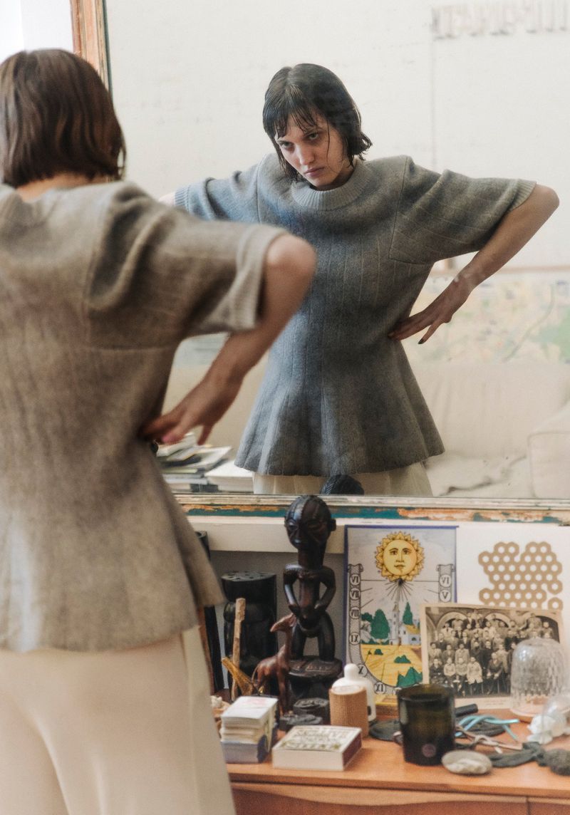 Adult woman in a gray short-sleeved, empire-waist dress with hands on her hips, posing in front of a mirror on a vanity.