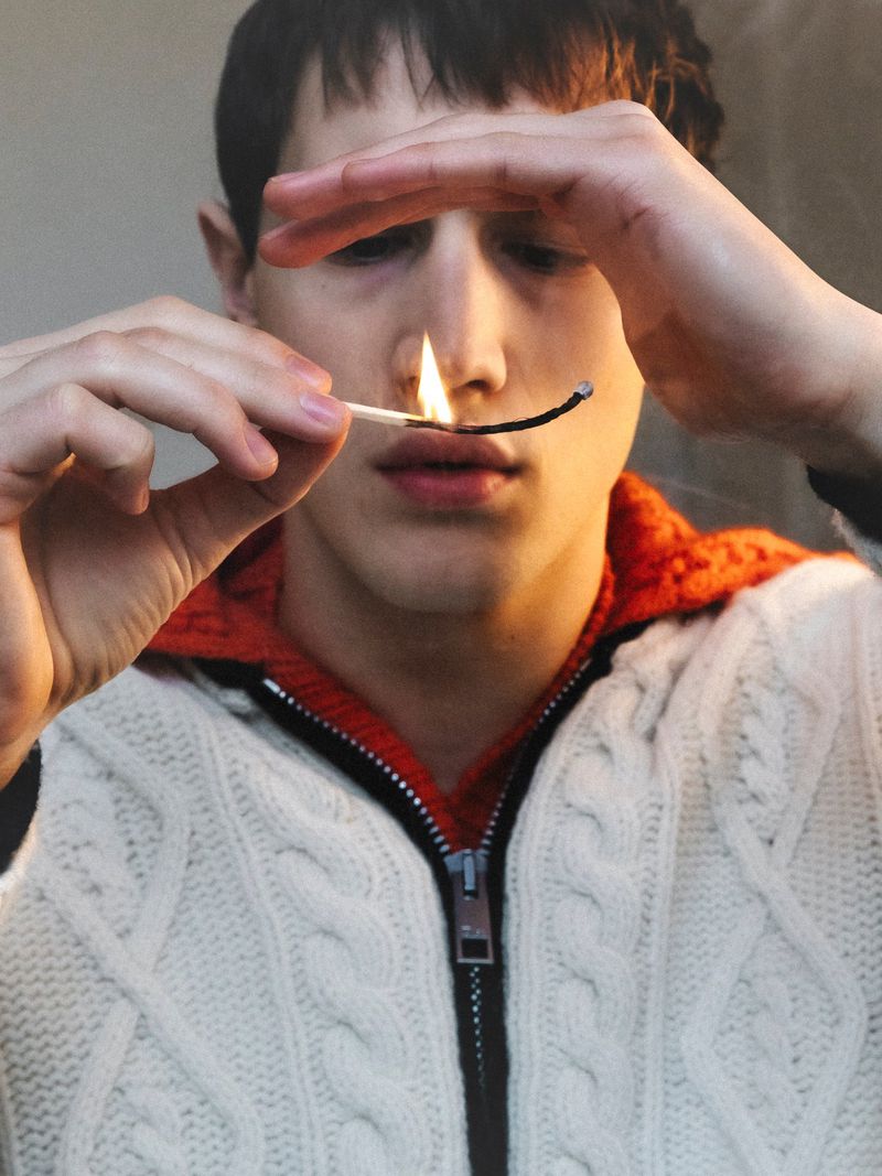 Close-up of a young adult man holding a lit match in front of his face, wearing a zipped, patterned sweater against a neutral backdrop.