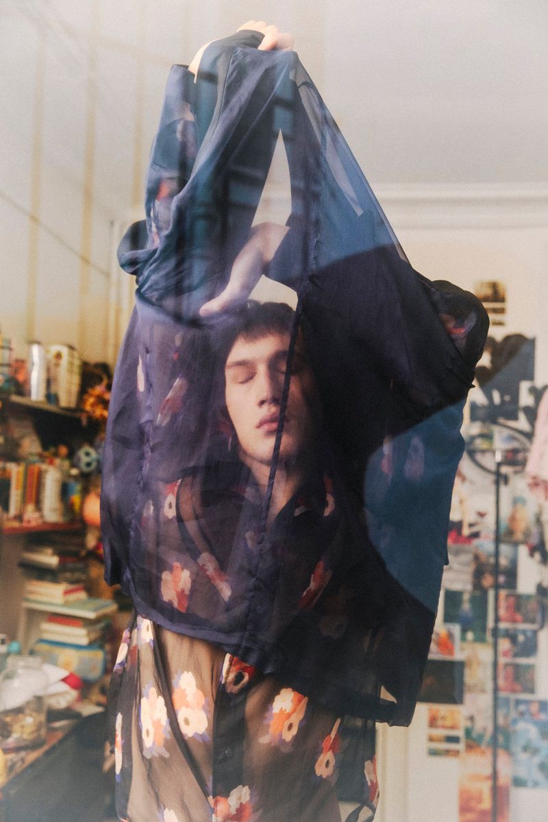 A young adult male poses indoors wearing a sheer floral-patterned shirt with a second floral shirt draped over his head and shoulders.
