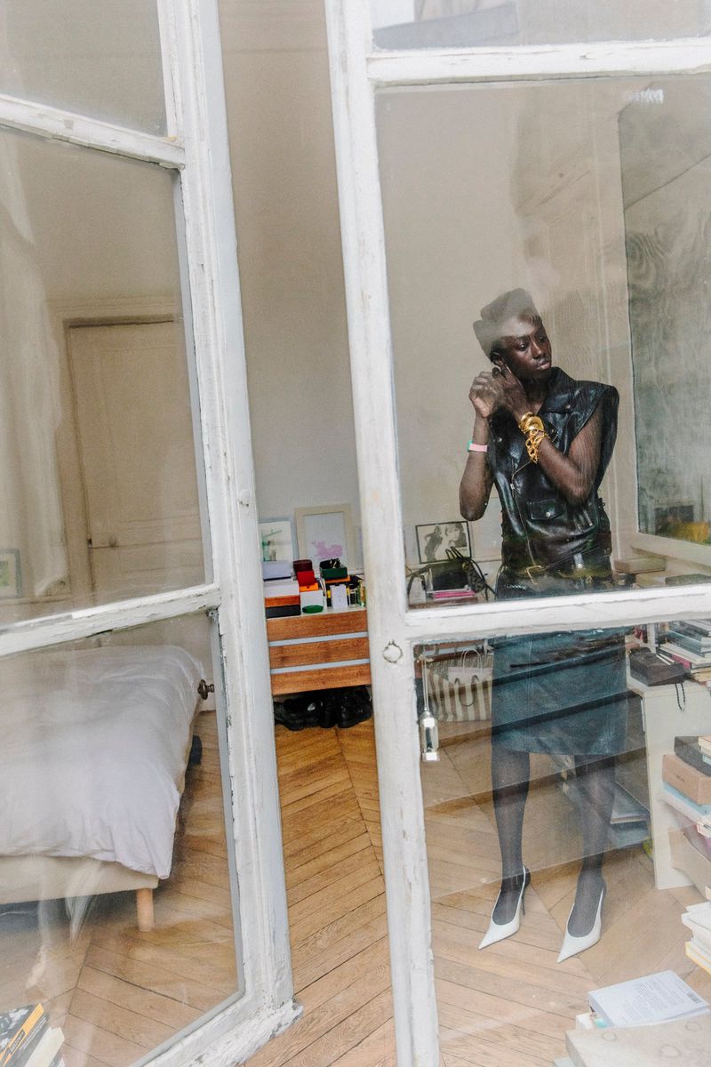 A Black woman in a black leather vest and skirt is seen in a room putting on an earring, reflected in a large glass window.