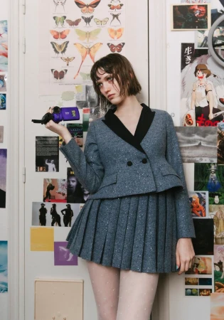 Woman in a blue blazer and pleated skirt stands in front of a white door with colorful posters and holds a vintage video camera.
