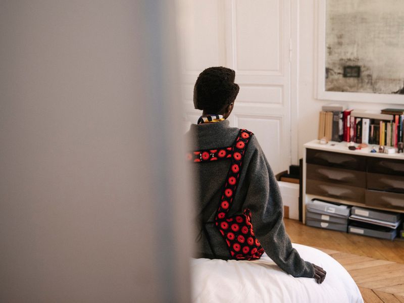 Back view of an African-American adult female in a gray sweater, patterned red bag, and white pants sitting on a bed indoors.