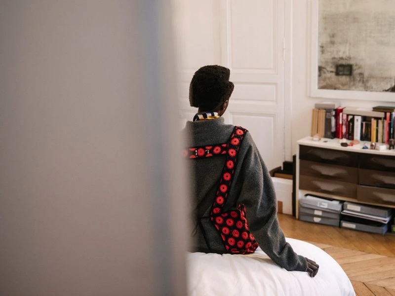 Back view of an African-American adult female in a gray sweater, patterned red bag, and white pants sitting on a bed indoors.