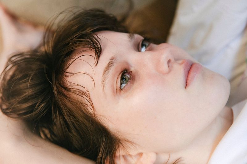 Close-up portrait of a young fair-skinned woman with brown hair lying down, looking up with a contemplative expression in a bright indoor setting.