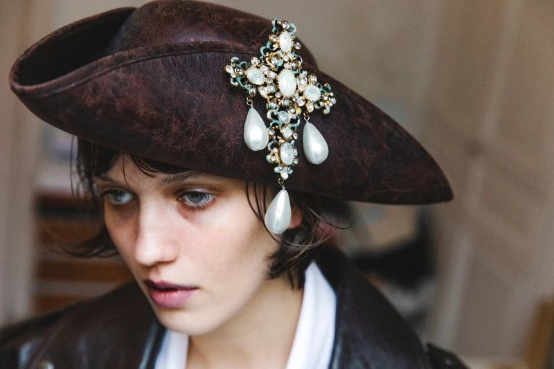 Adult woman in a brown hat with a pearl cross earring, looking down with a neutral facial expression, wearing a leather jacket.