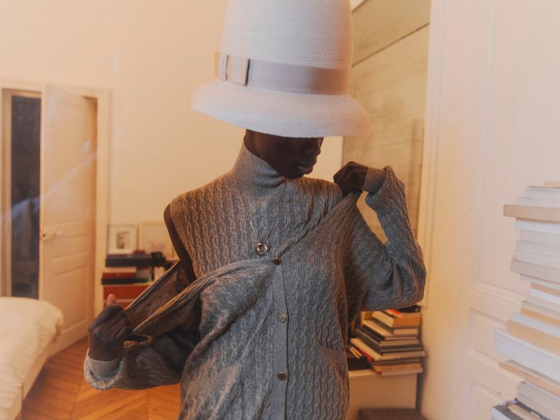 Indoor portrait of a black female adult wearing a large white hat and grey patterned cardigan with piles of books in the background.