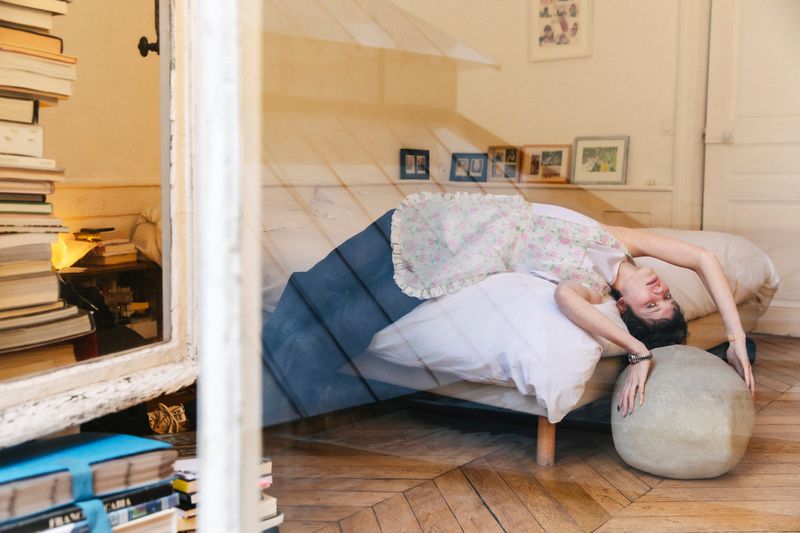 Woman in a floral dress lies across a bed with a white pillow and blue blanket, next to a stack of books in an indoor bedroom setting.