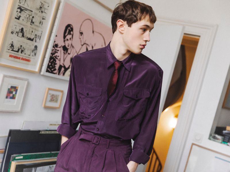 Young adult man in a purple suit and tie stands in a room with framed artwork on the walls, creating a sophisticated and fashionable look.