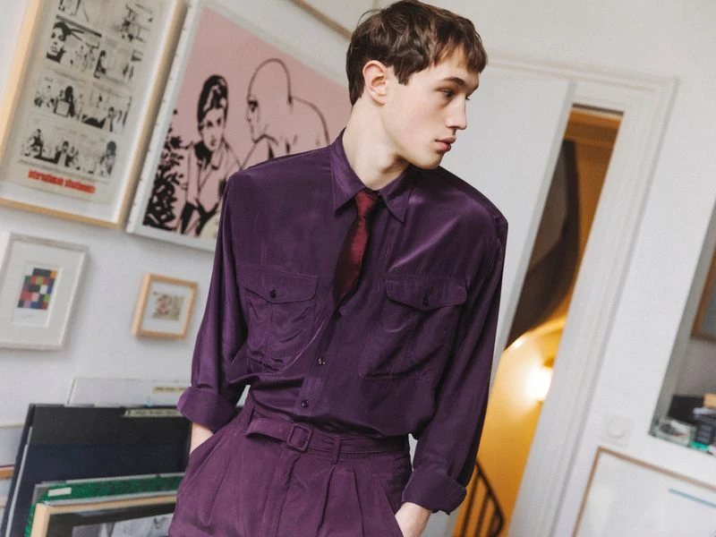 Young adult man in a purple suit and tie stands in a room with framed artwork on the walls, creating a sophisticated and fashionable look.