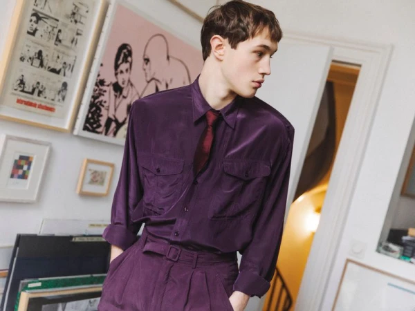 Young adult man in a purple suit and tie stands in a room with framed artwork on the walls, creating a sophisticated and fashionable look.
