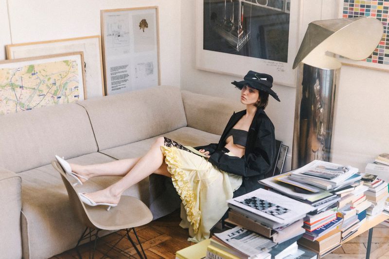 Woman in black jacket and hat relaxes on beige sofa near a table covered in books in a bright, art-filled room with natural light.