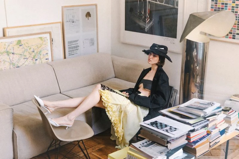 Woman in black jacket and hat relaxes on beige sofa near a table covered in books in a bright, art-filled room with natural light.