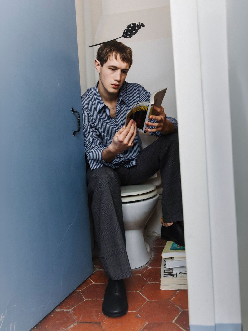 Man reading a magazine while sitting on a toilet with books underneath in a small bathroom with blue door and terra cotta floors.
