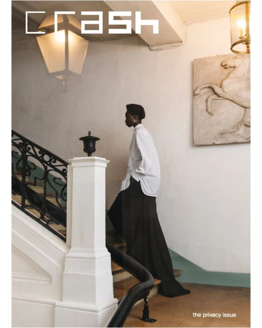 A person walks down stairs in a fashion editorial shot inside a building with white walls, a black metal railing, and light fixtures.