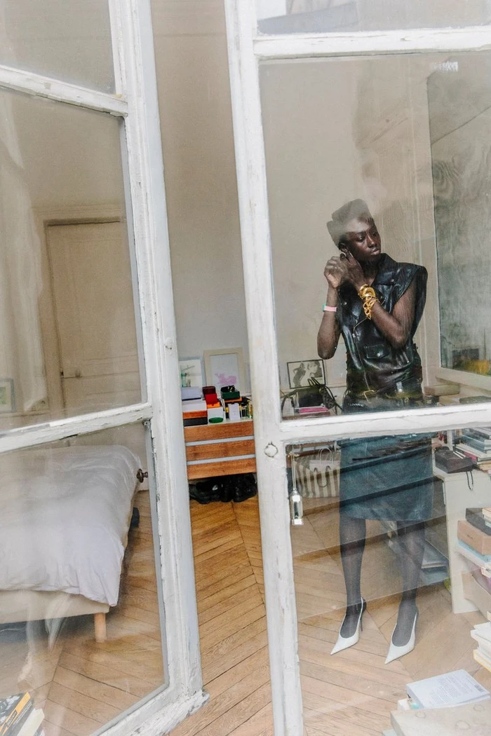 Black woman wearing a dark sleeveless top and white heels, viewed through a framed glass door in a bedroom setting.