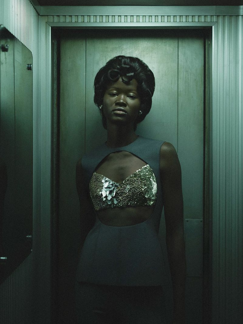 Glamorous portrait of a Black woman in a blue dress with a cut-out and ornate, shiny fabric, posing in a textured elevator.