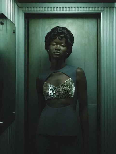 Glamorous portrait of a Black woman in a blue dress with a cut-out and ornate, shiny fabric, posing in a textured elevator.
