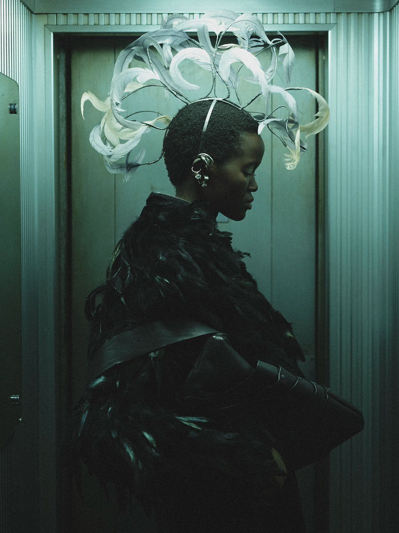Fashionable woman in feathered dress and extravagant feathered headpiece poses in metallic elevator with a side profile view.
