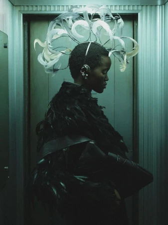 Fashionable woman in feathered dress and extravagant feathered headpiece poses in metallic elevator with a side profile view.